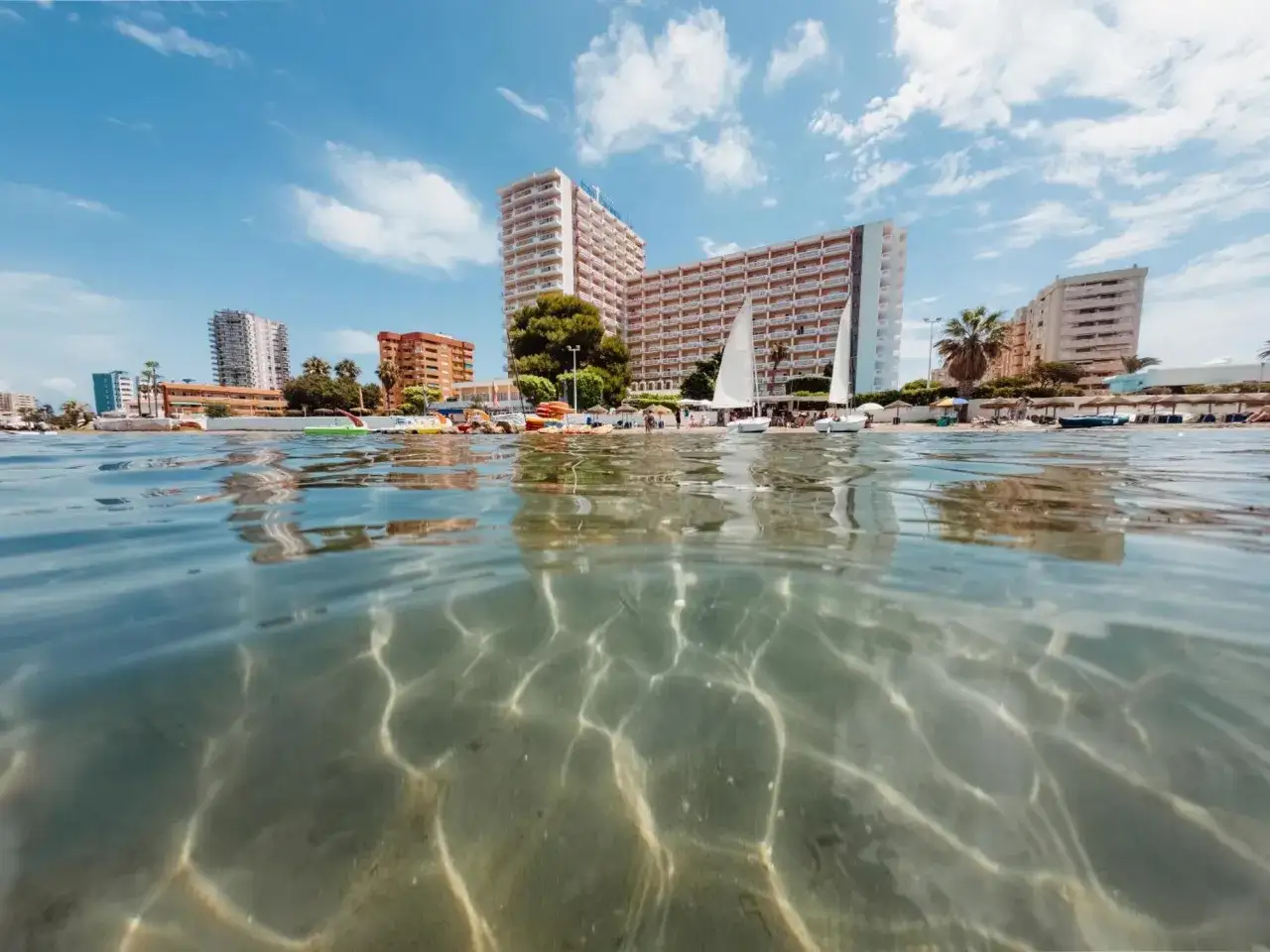 Widok z wody na plażę w La Manga z hotelami i żaglówkami. Słońce tworzy wzory na piaszczystym dnie.