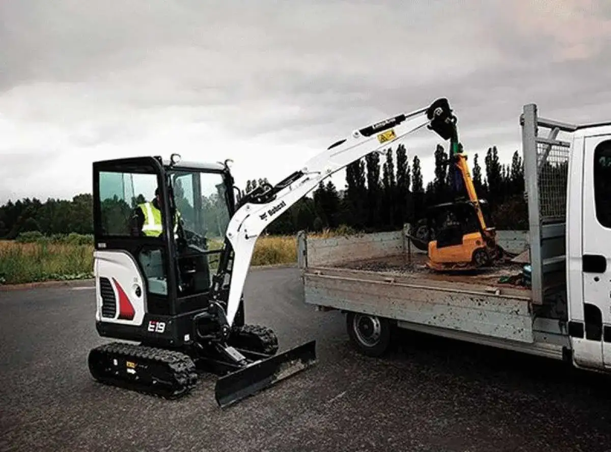 Koparka gąsienicowa Bobcat E19, z operatorem w kamizelce, ładuje mniejszą maszynę na przyczepę. Dane techniczne Bobcat E19 są imponujące.