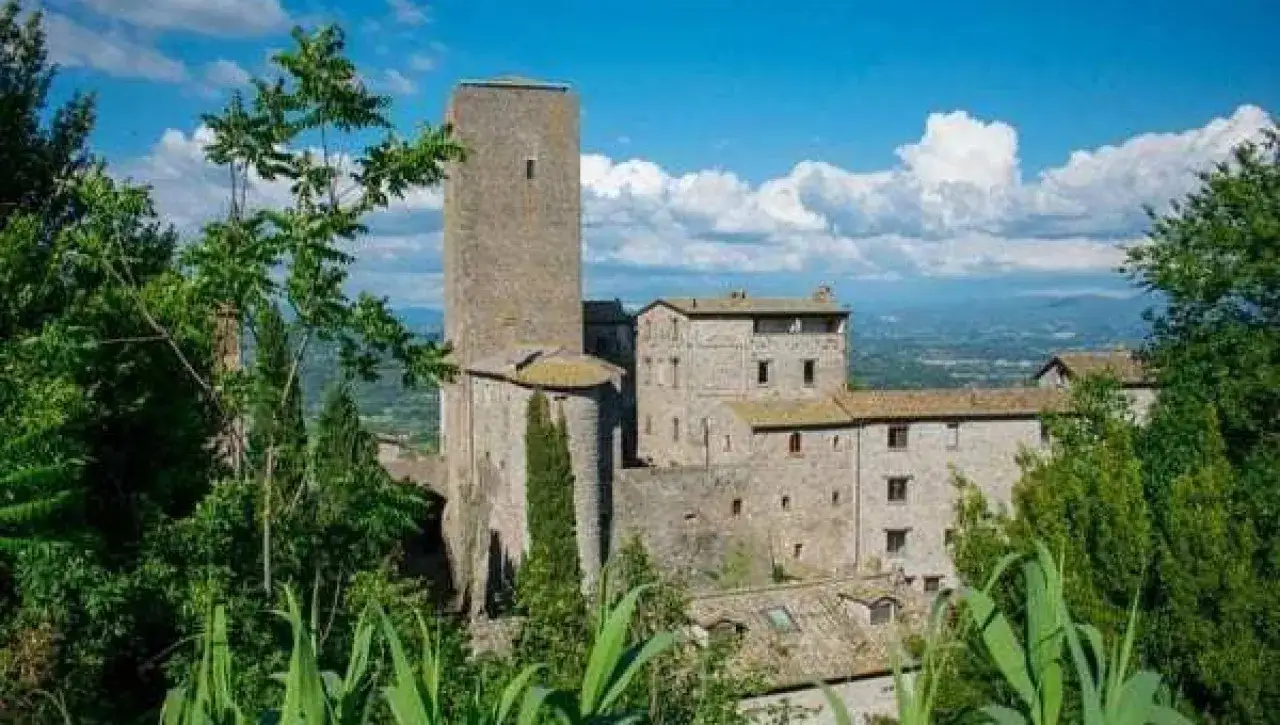 Antico borgo fortificato di Bassano in Teverina, con torre imponente e case in pietra, immerso nel verde sotto un cielo azzurro con nuvole bianche.