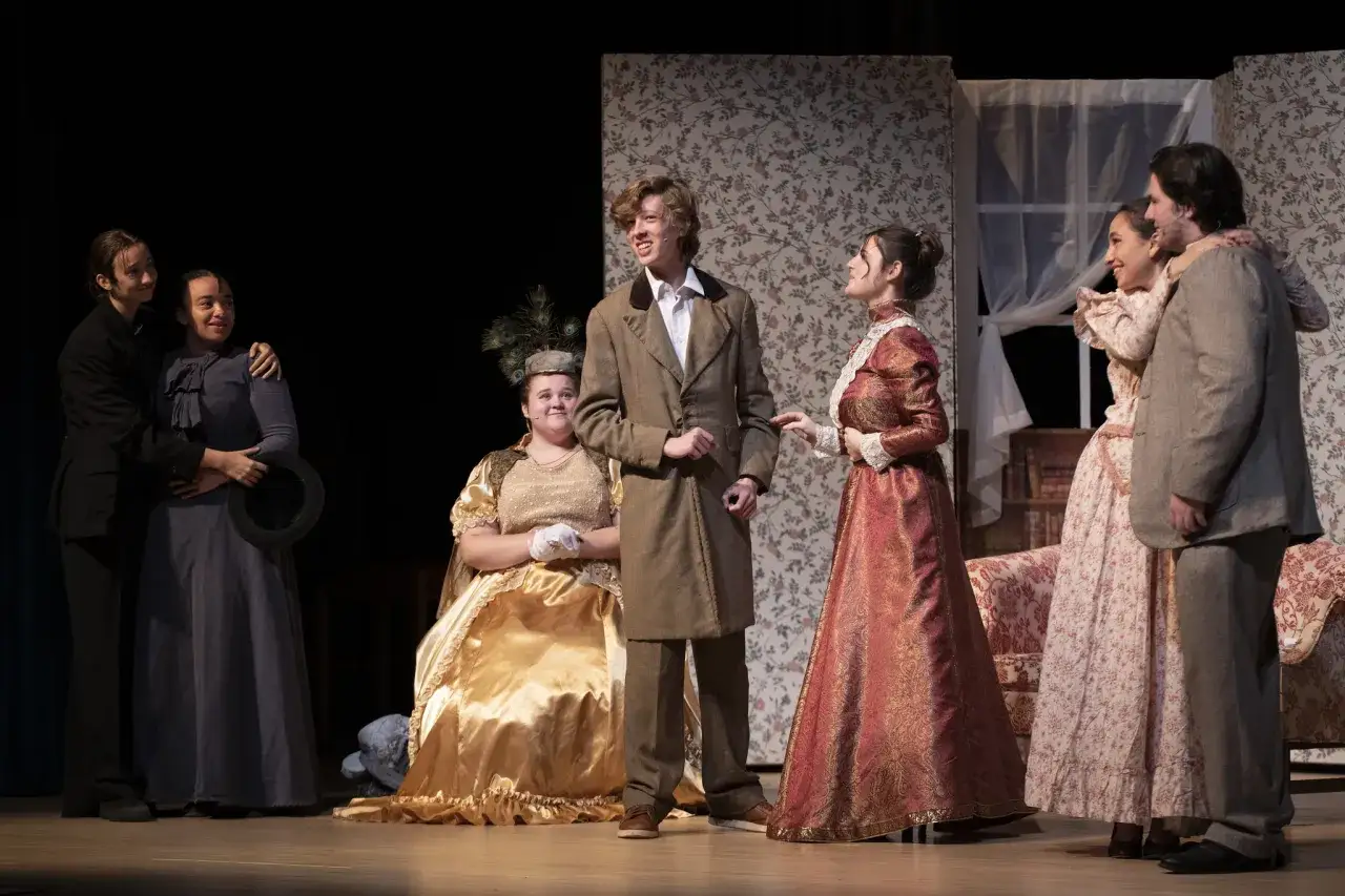 Skådespelare i historiska dräkter på scenen. En kvinna i guldklänning med påfågelsfjädrar i håret, en man i brun kavaj och två par som omfamnar varandra. Scenografi med blommig tapet och möbler. Rollistan i pelikanfallet.