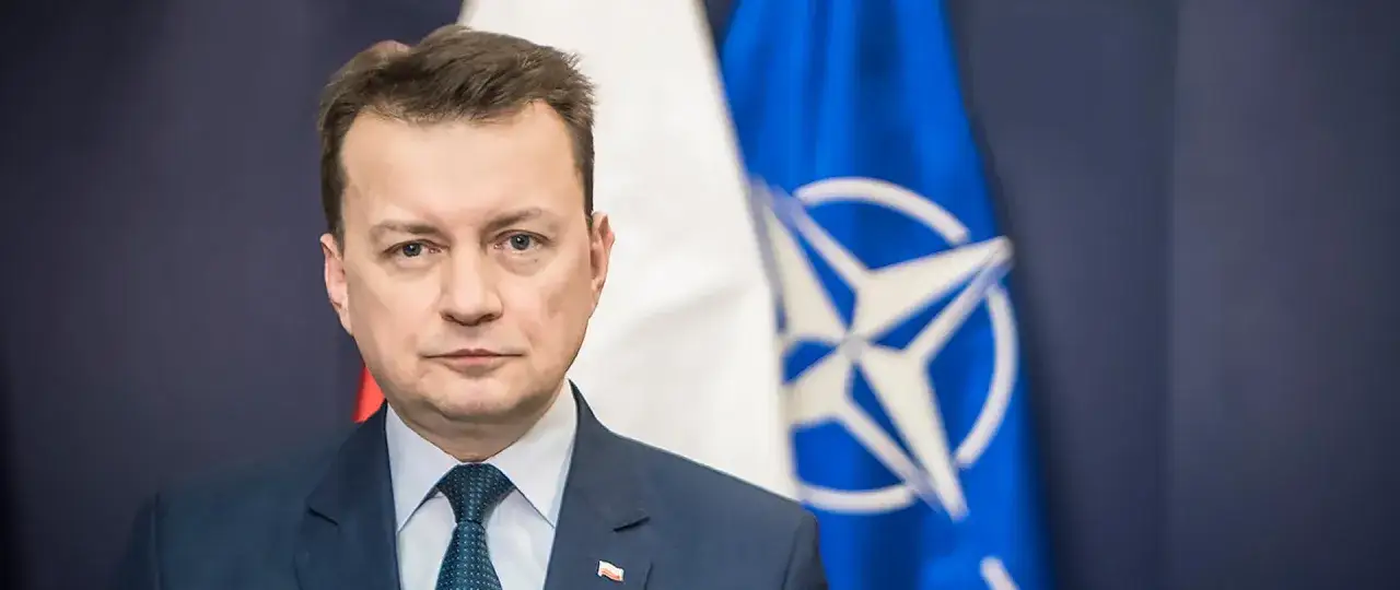 Minister obrony narodowej w garniturze, na tle flagi Polski i NATO.