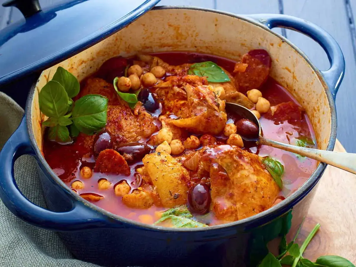 Un plat mijoté de poulet chorizo, pois chiches et olives noires dans une cocotte bleue. Des herbes fraîches apportent une touche de couleur.