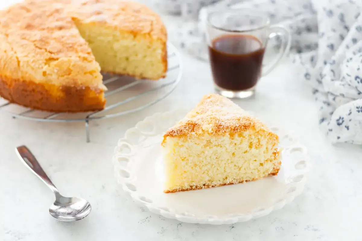 Una fetta di torta ricotta e cocco, soffice e dorata, su un piatto bianco. Accanto, una tazzina di caff&egrave; e la torta intera su una griglia.