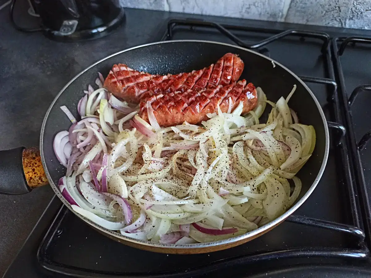 Jak usmażyć kiełbasę na patelni? Dwie kiełbaski skwierczą na patelni z cebulą, posypane pieprzem.