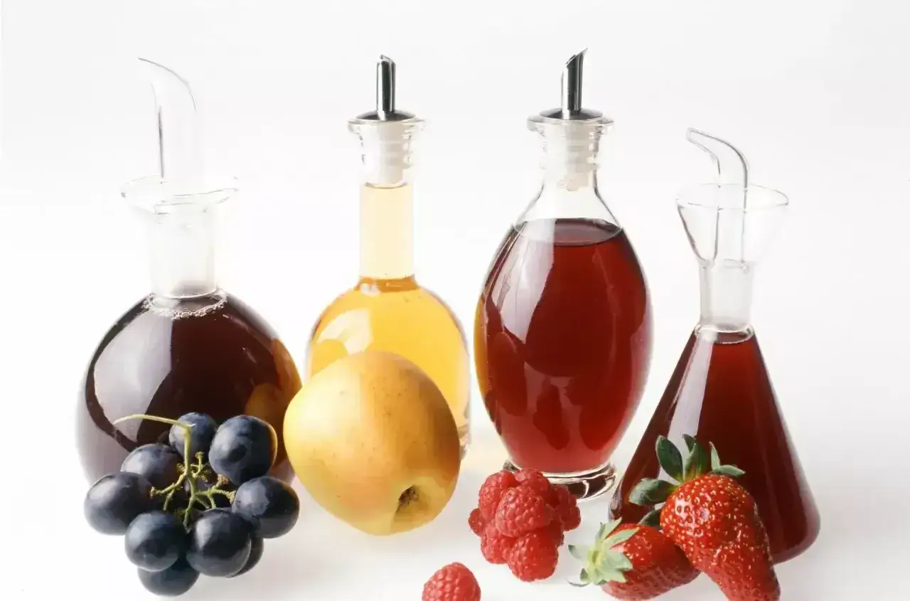 Butelki z octem, w tym ocet balsamiczny malinowy do czego? Obok owoce: winogrona, jabłko, maliny i truskawki.