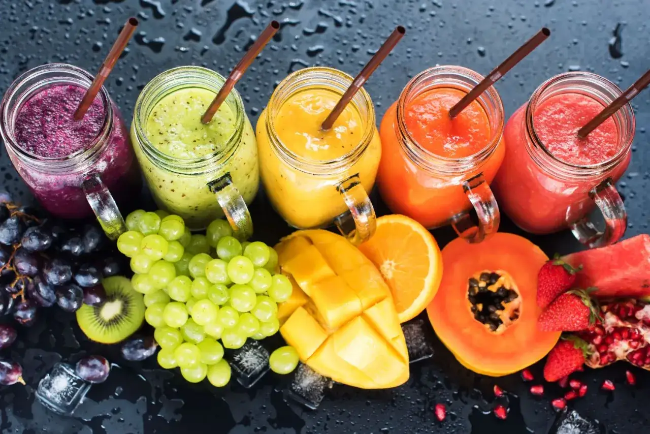 Kolorowe smoothie w słoikach, obok świeże owoce: winogrona, mango, papaja, pomarańcza, truskawki, arbuz i granat. Pyszny kordiał lata.