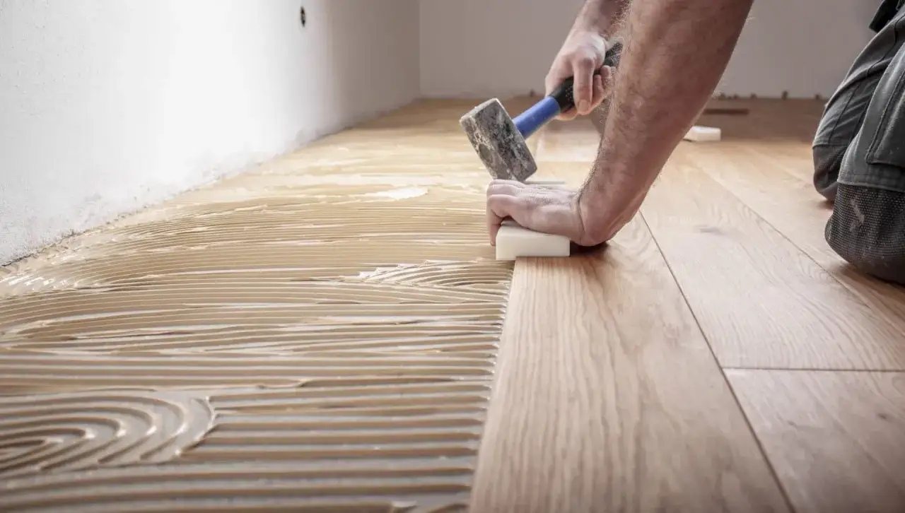 Pose d'un parquet en bois, avec de la colle appliqu&eacute;e au sol. Un artisan utilise un maillet pour ajuster les lames. Travaux pour refaire sol maison.