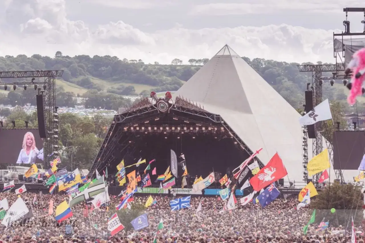 A massive crowd gathers before the iconic Glastonbury Pyramid Stage, waving flags under a cloudy sky. Dolly Parton smiles from a large screen.