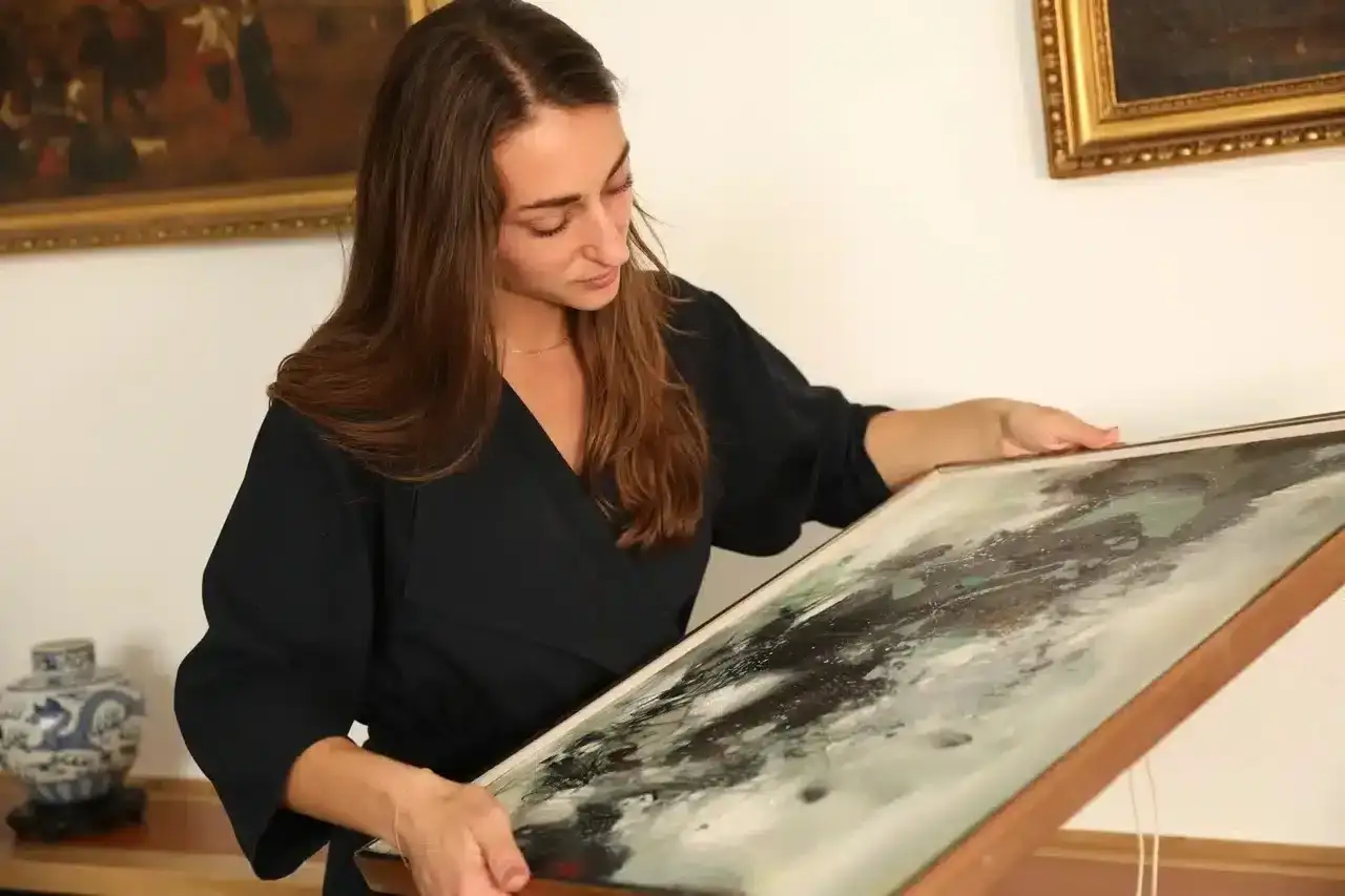 Une femme examine attentivement un tableau abstrait, ses mains tenant le cadre. Elle semble expertiser un tableau.