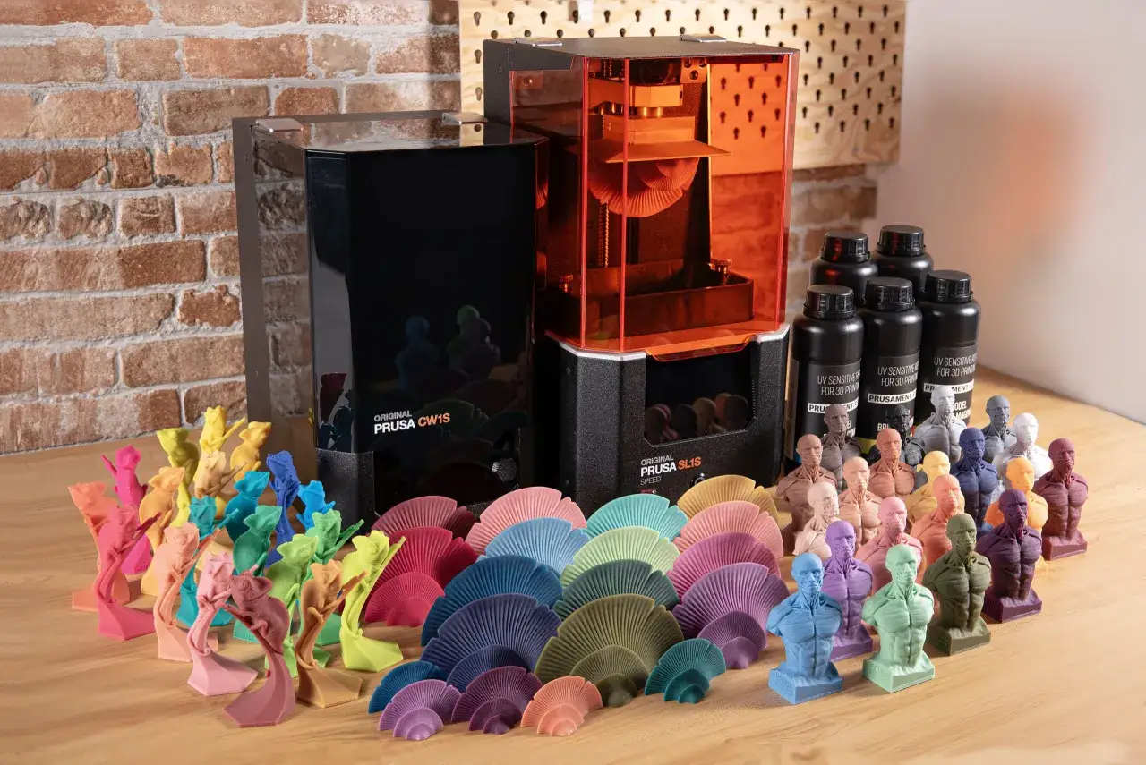 A color resin 3D printer sits beside bottles of resin and a vibrant collection of 3D printed figures, including chameleons and anatomical busts.