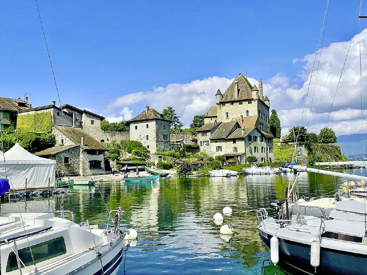 Vue d'une aire de camping-car au bord du lac Léman, avec un château médiéval, des bateaux et des maisons en pierre.