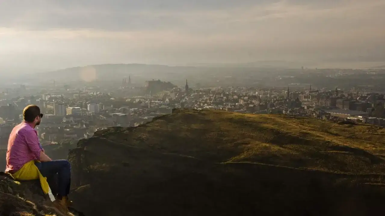 Mann sitzt auf einem H&uuml;gel und blickt auf Edinburgh im Oktober. Die Stadt erstreckt sich unter ihm, mit dem Schloss in der Ferne.