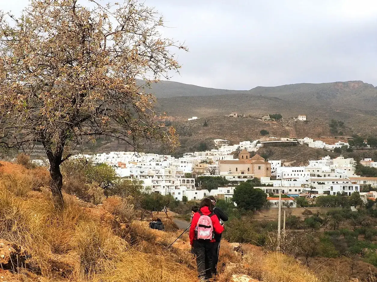 Senderistas caminan por un sendero seco hacia los pradillos, un pueblo blanco enclavado en una ladera monta&ntilde;osa.