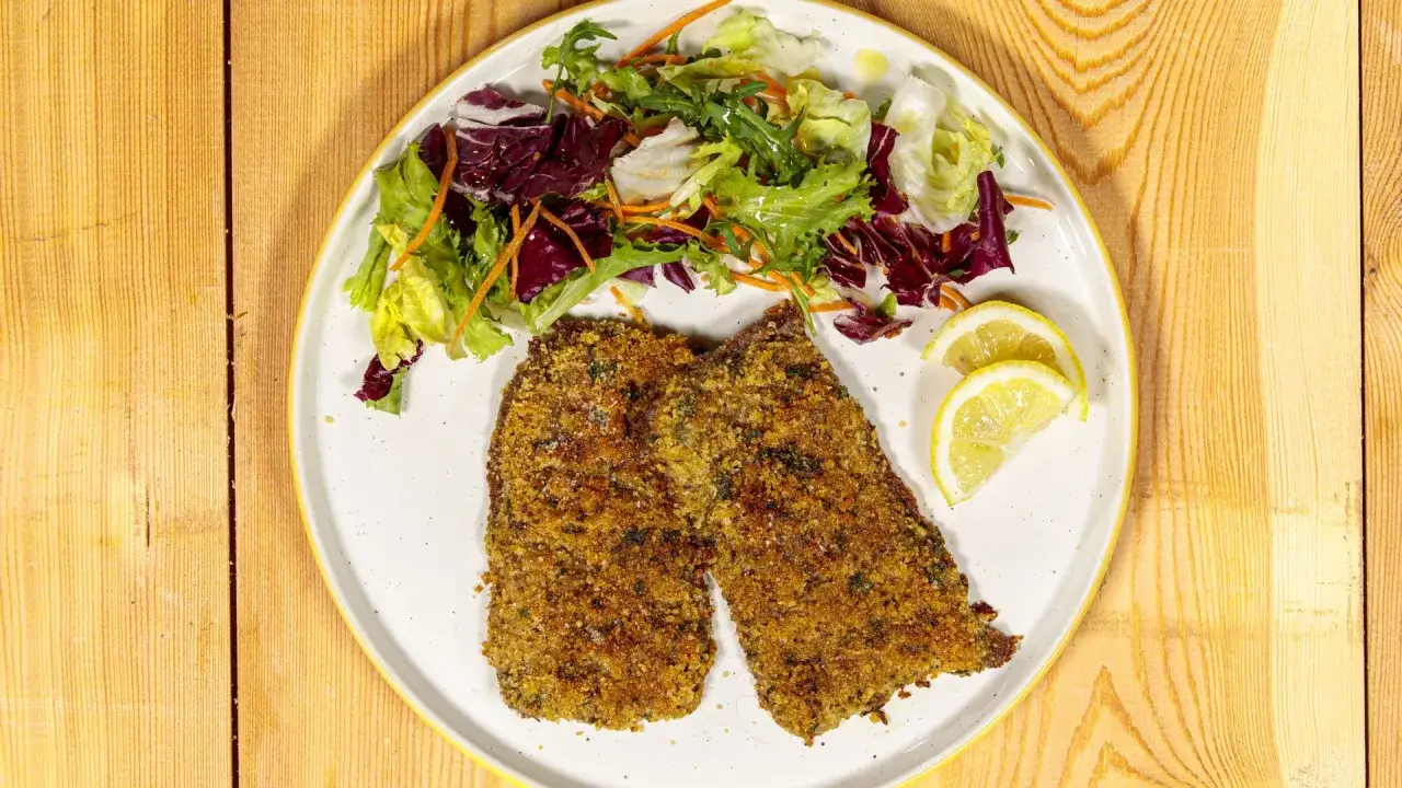 Due cotolette alla palermitana dorate e croccanti, servite con un'insalata fresca e spicchi di limone su un piatto bianco.
