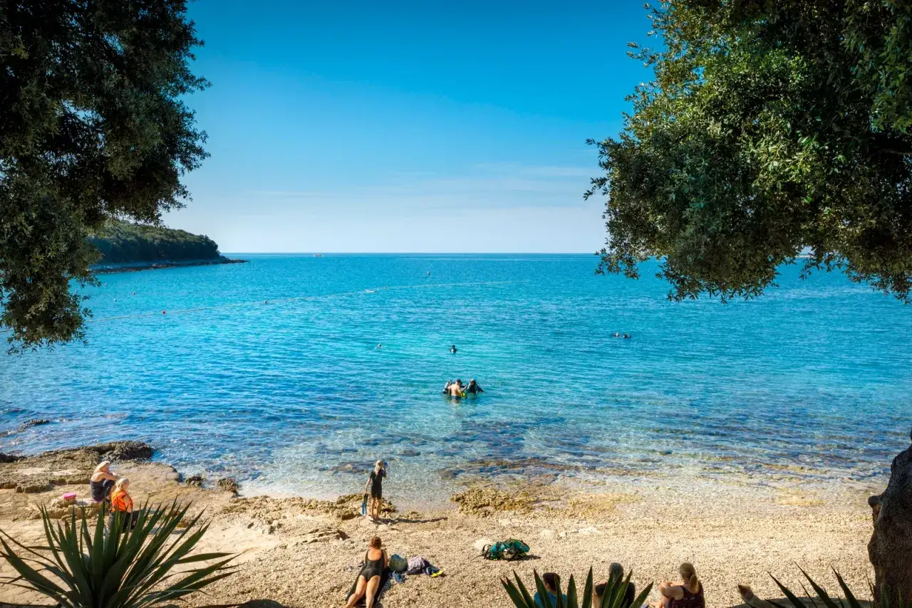 Słoneczny dzień na plaży w Porto Sole. Ludzie odpoczywają na kamienistym brzegu, inni pływają i nurkują w krystalicznie czystej wodzie. Idealne miejsce na camping.