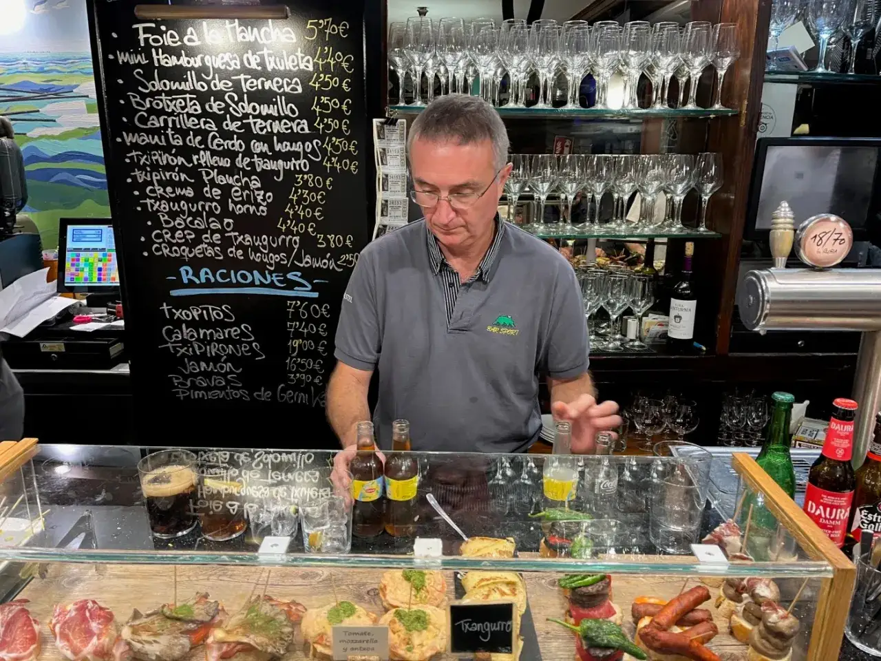 Man vid en bardisk i San Sebasti&aacute;n, med en meny med tapas och raciones p&aring; svarta tavlan.