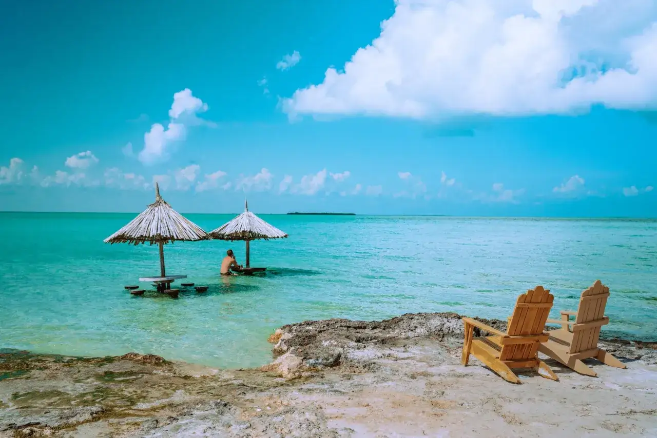 Idealne belize wakacje: dwa drewniane leżaki na plaży, a w turkusowej wodzie stolik pod parasolem, przy którym siedzi mężczyzna.
