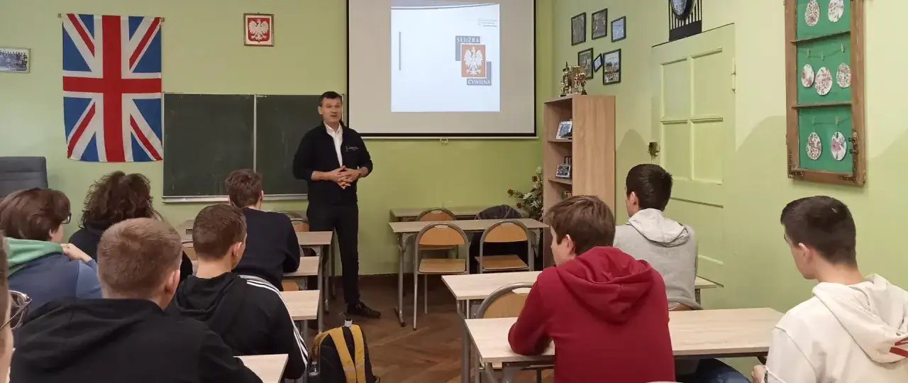 Nauczyciel prowadzi prezentację dla uczniów. Na ścianie flaga Wielkiej Brytanii i godło Polski. To może być przygotowanie do lekcji lub spotkanie rady pedagogicznej.