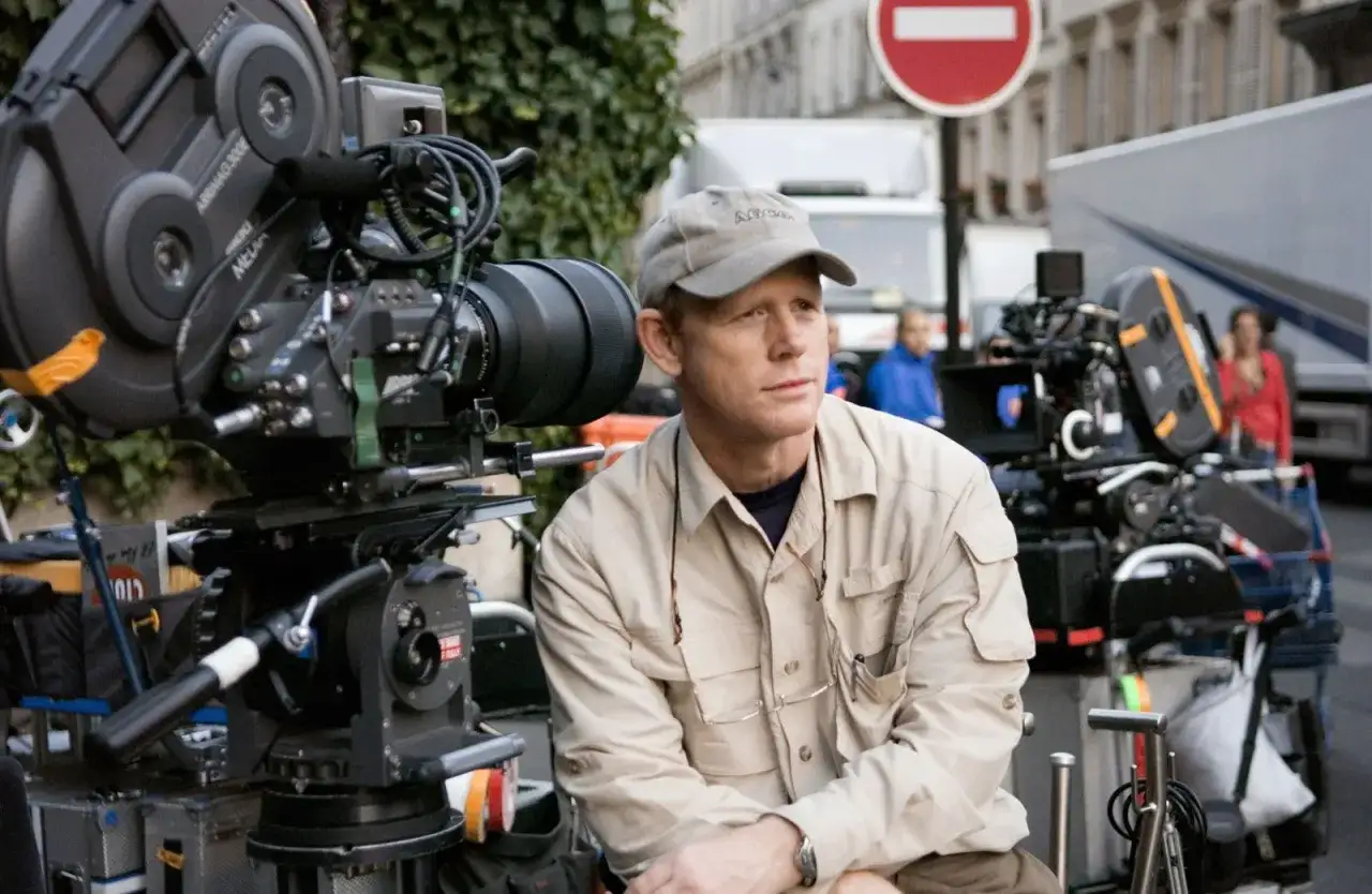 Ron Howard, med keps och beige skjorta, sitter vid en filmkamera på en inspelningsplats.