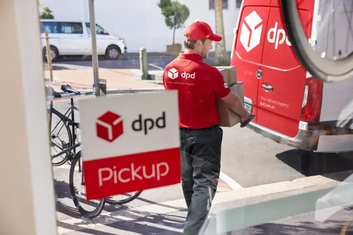 Kurier DPD w czerwonej koszulce i czapce niesie paczki do czerwonego vana. Obok widoczny znak DPD Pickup.