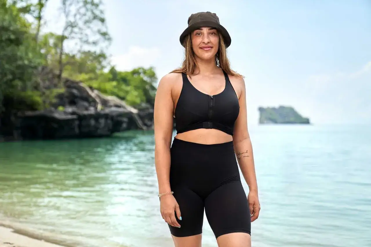 Sandra Cucarano på en strand med gröna öar i bakgrunden. Hon bär en svart bikini och en hatt.