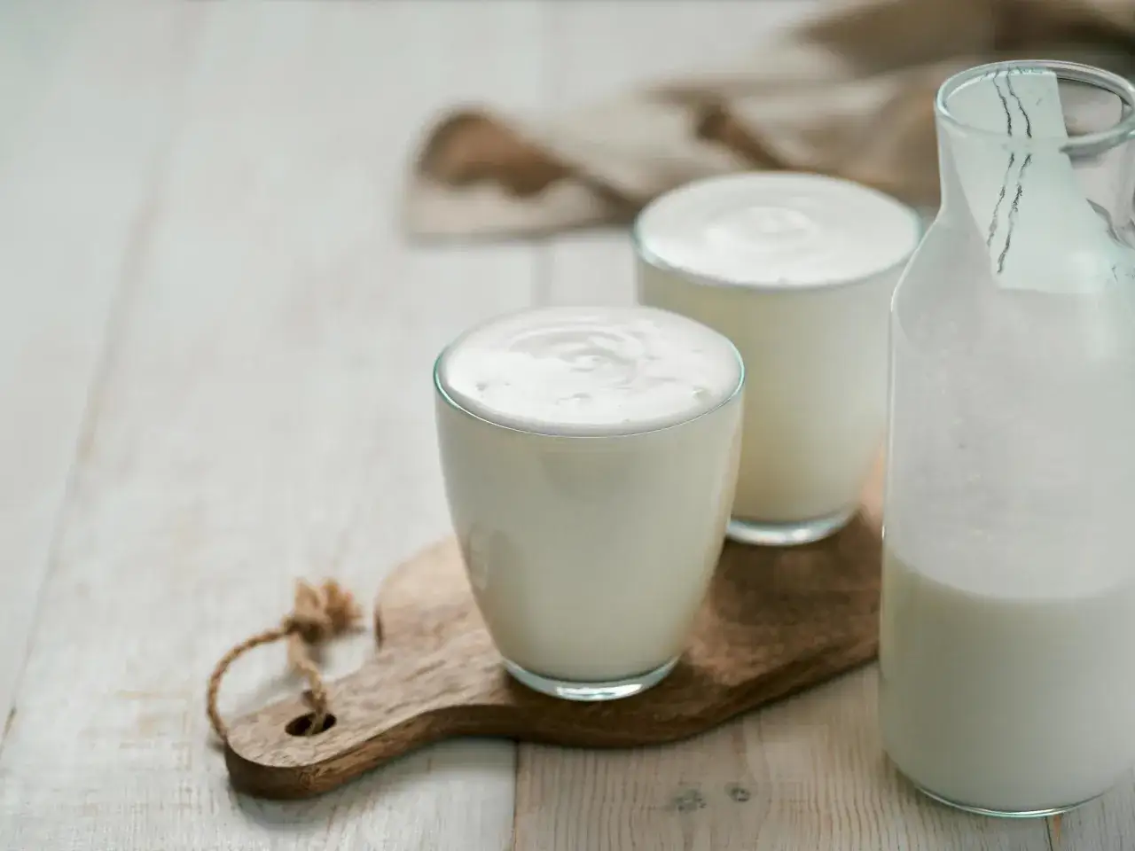 Erfrischender Ayran, perfekt zum selber machen. Zwei Gl&auml;ser und eine Flasche gef&uuml;llt mit dem cremigen Getr&auml;nk auf einem Holzbrett.
