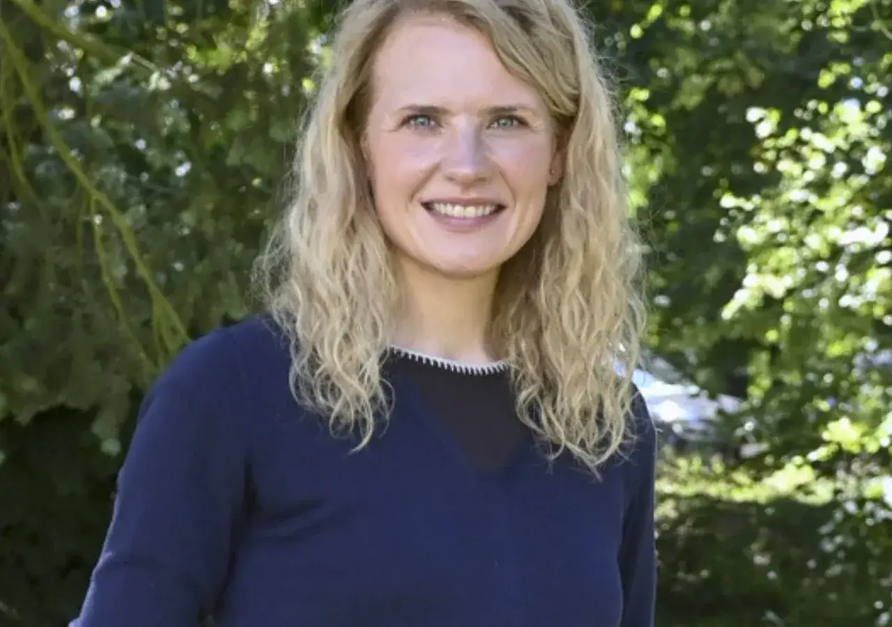Uśmiechnięta Wiktoria Grodecka z kręconymi blond włosami, w granatowym swetrze, na tle zieleni.