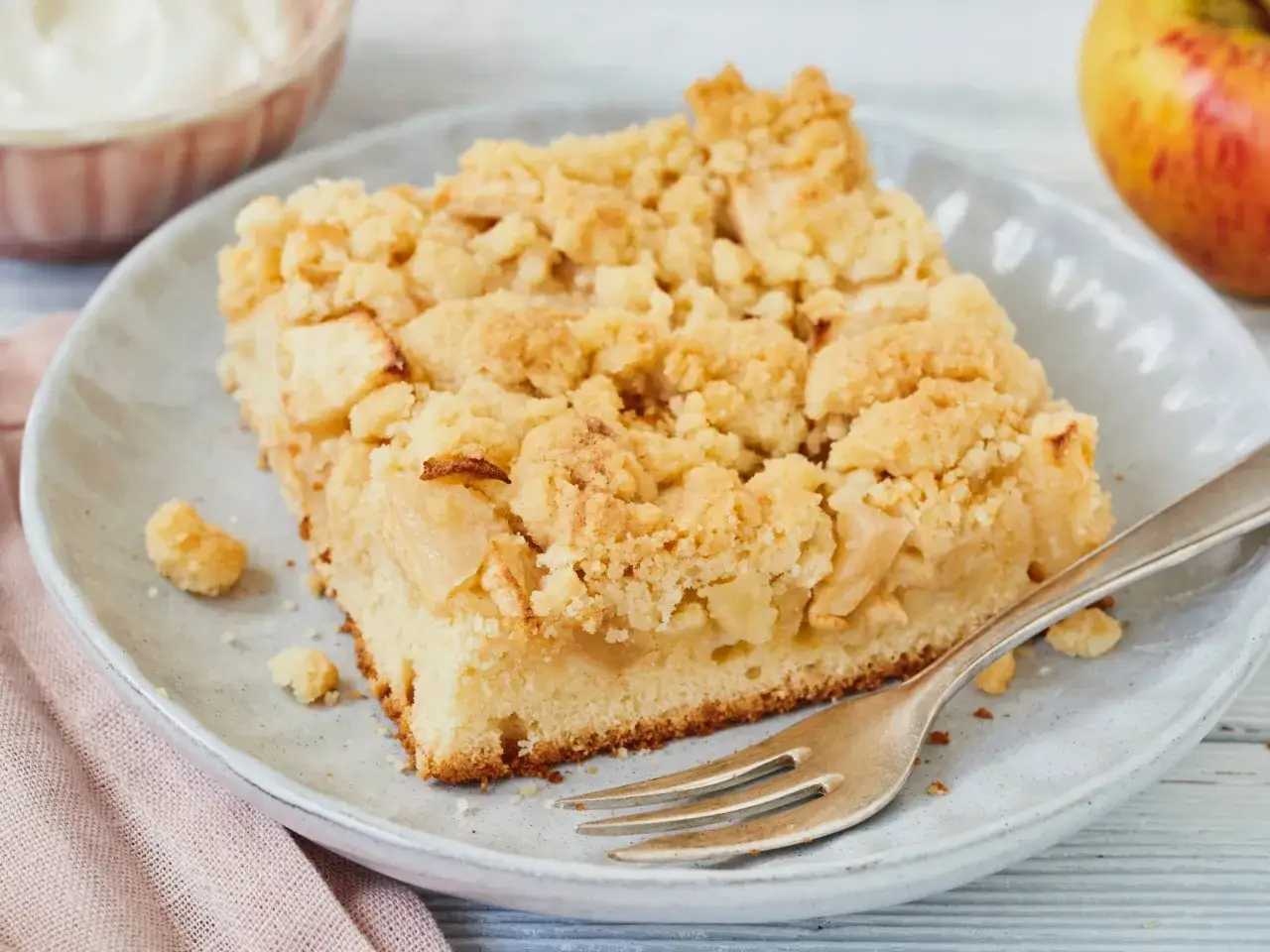 Ein Stück Apfelkuchen mit Streuseln auf einem Teller, wie er in vielen **Landfrauen Kuchen Rezepte** zu finden ist. Dazu eine Gabel und ein Apfel.