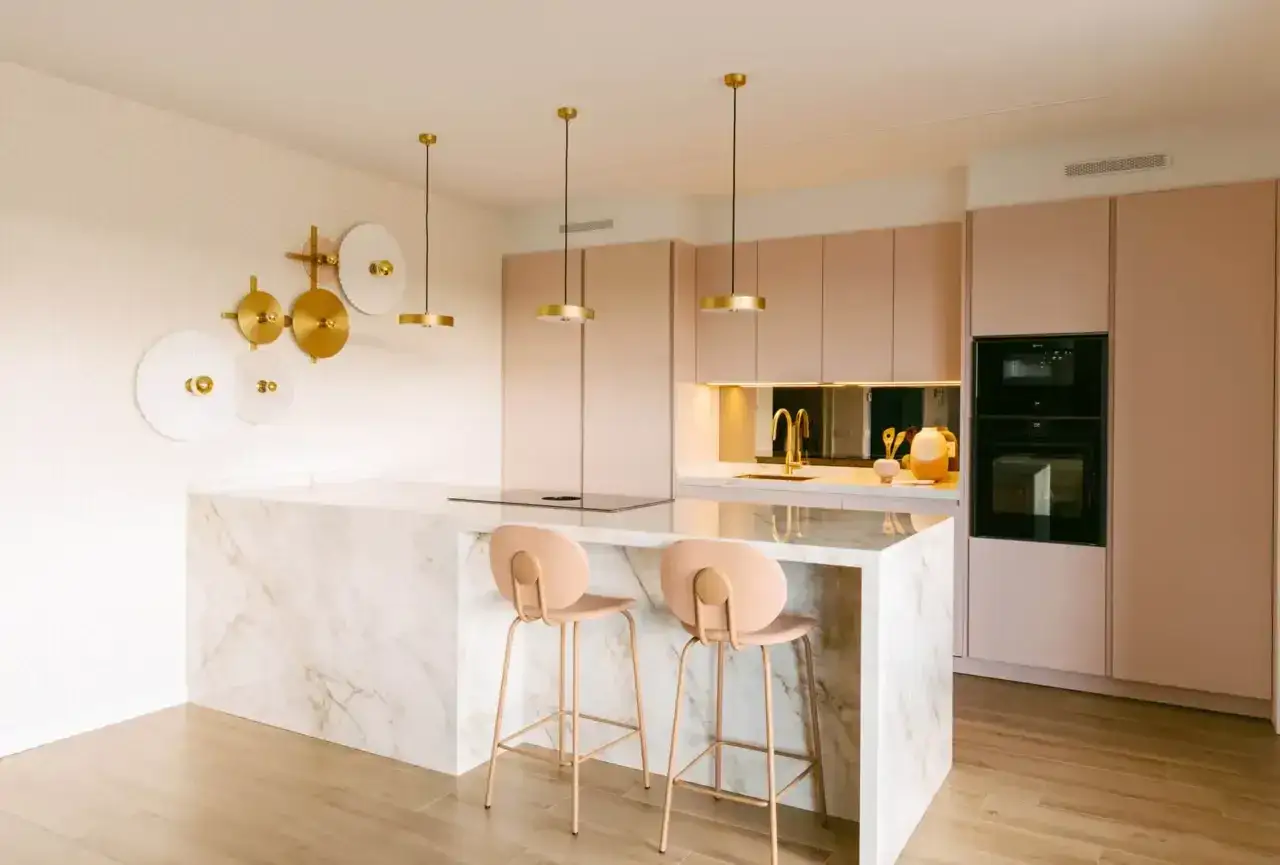 Elegante cocina rosa con isla de mármol, taburetes a juego y detalles dorados. Un espacio moderno y acogedor.