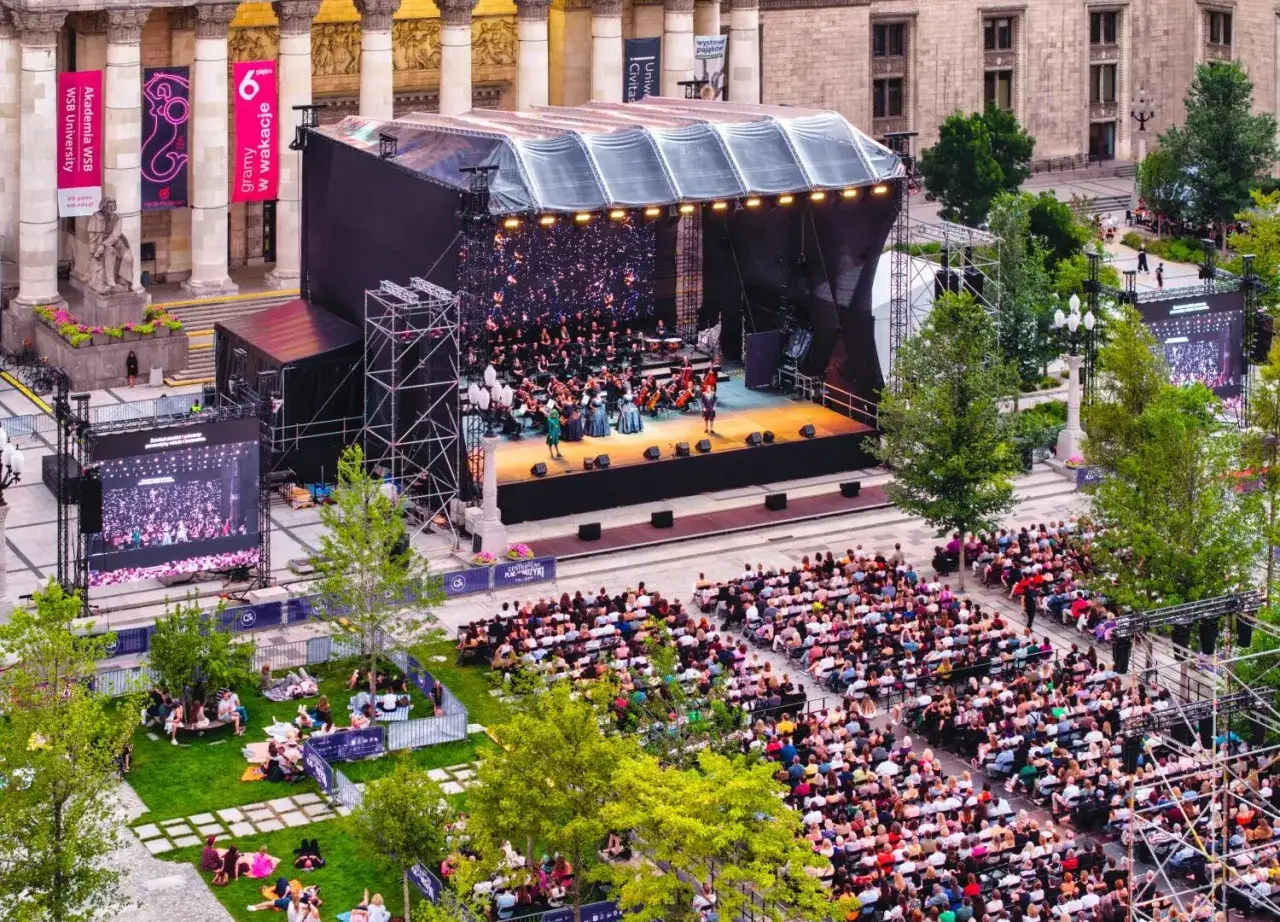 Koncert plenerowy w Warszawie. Orkiestra i soliści na scenie, publiczność siedzi na krzesłach i trawie. Wspaniałe warszawskie wydarzenia kulturalne.