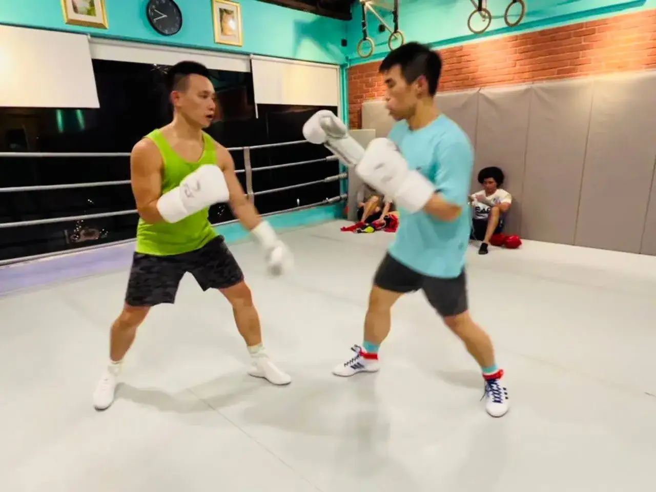 Two men are engaged in a boxing match in a gym. One man wears a green tank top and the other a blue t-shirt.