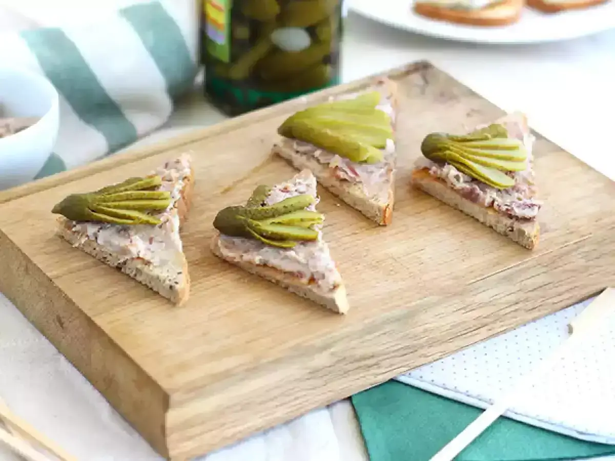 Quatre toasts triangulaires tartin&eacute;s de rillettes de canard, garnis de cornichons croquants, sur une planche en bois.