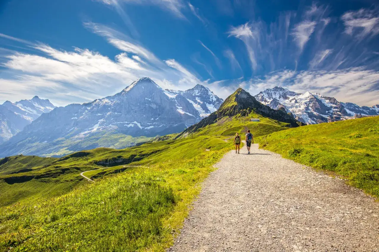 Two hikers enjoy a scenic path through the Swiss Alps, with majestic snow-capped mountains under a dramatic sky.