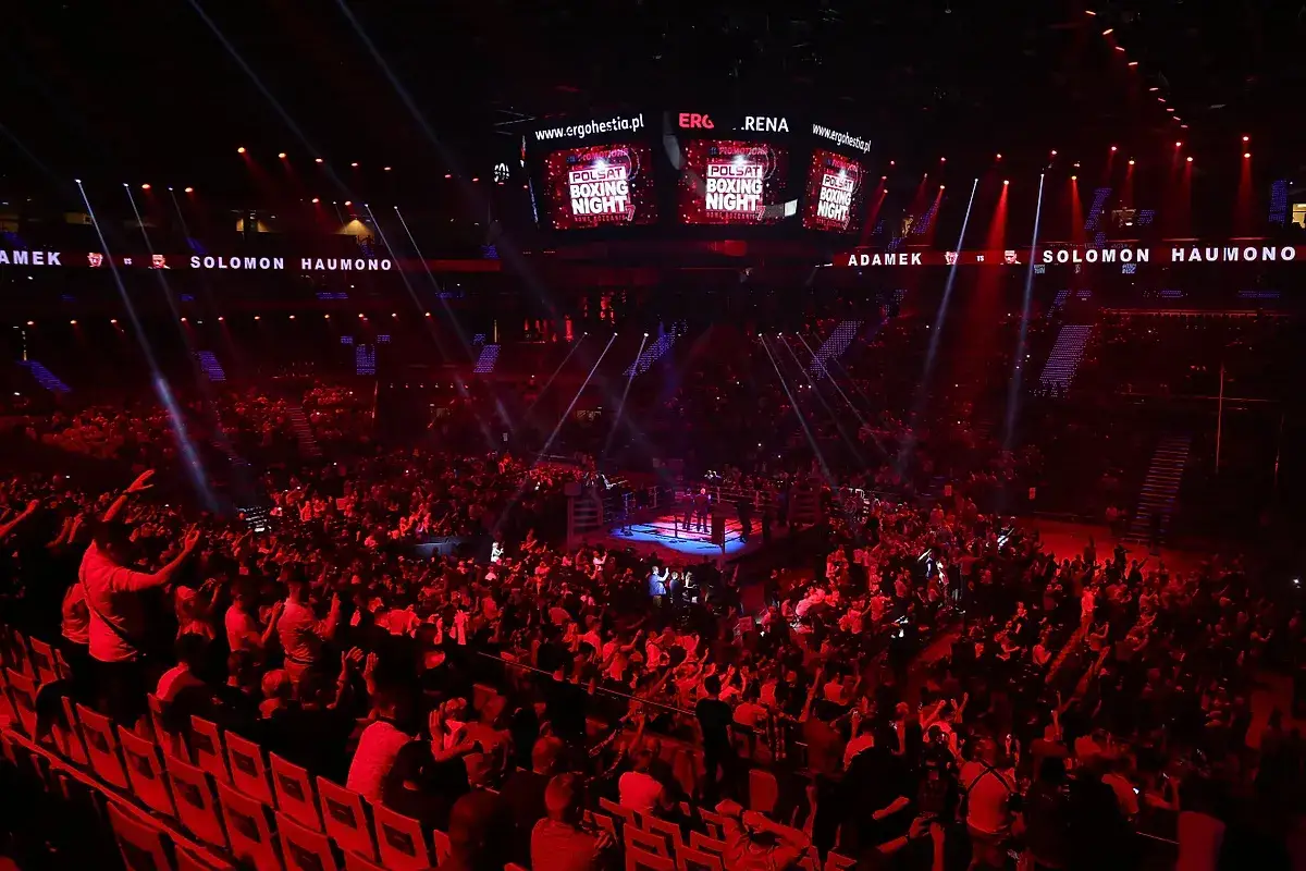 Widownia w czerwonym świetle podczas Polsat Boxing Night. Na ekranach nazwy zawodników i logo wydarzenia.