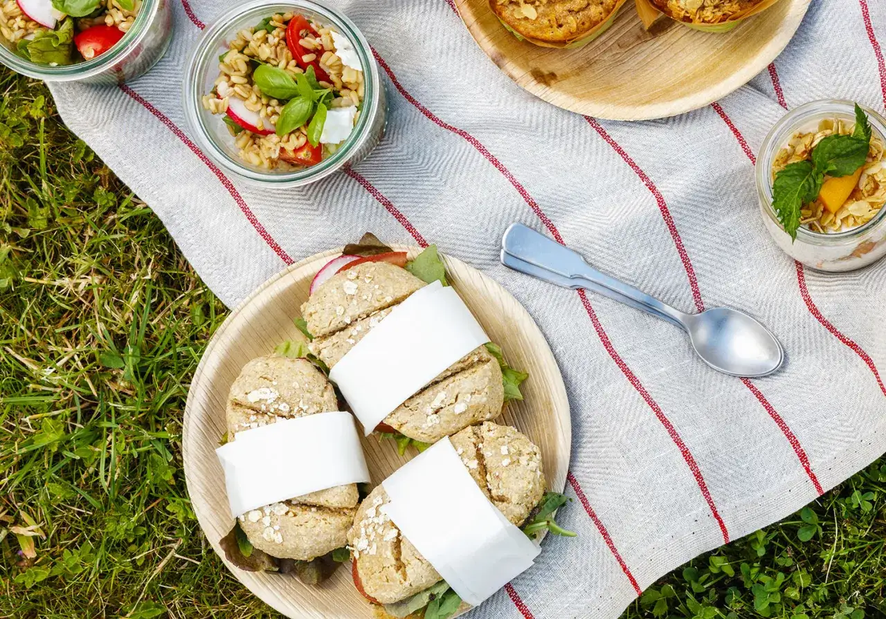 Une belle idée pique-nique : petits pains garnis, salades en bocaux et muesli sur une nappe à rayures rouges, posée sur l'herbe.