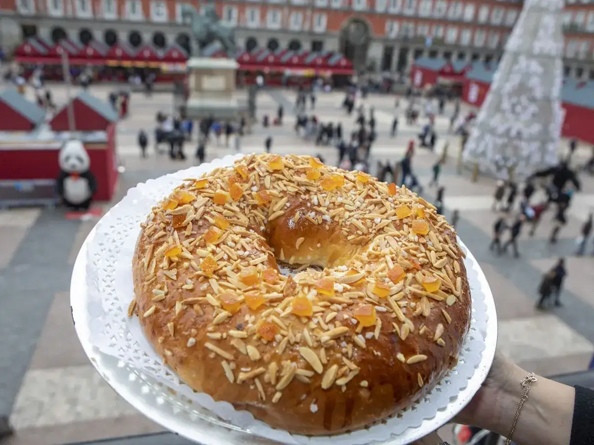 El mejor roscón de Madrid, adornado con almendras y fruta confitada, se presenta en la Plaza Mayor, con gente y puestos navideños al fondo.