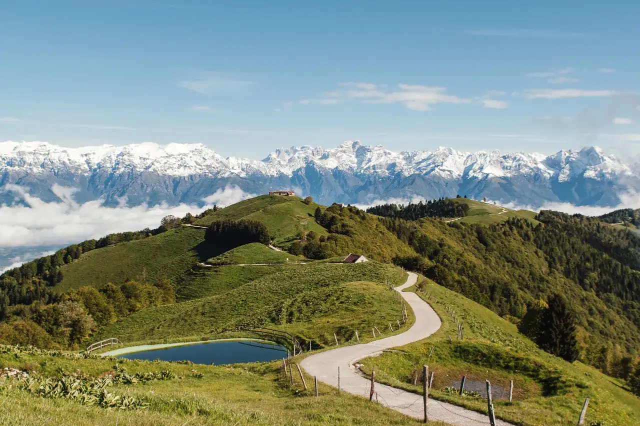 Malga Gardà Lentiai: sentiero serpeggiante tra verdi colline, laghetto alpino e maestose cime innevate all'orizzonte.