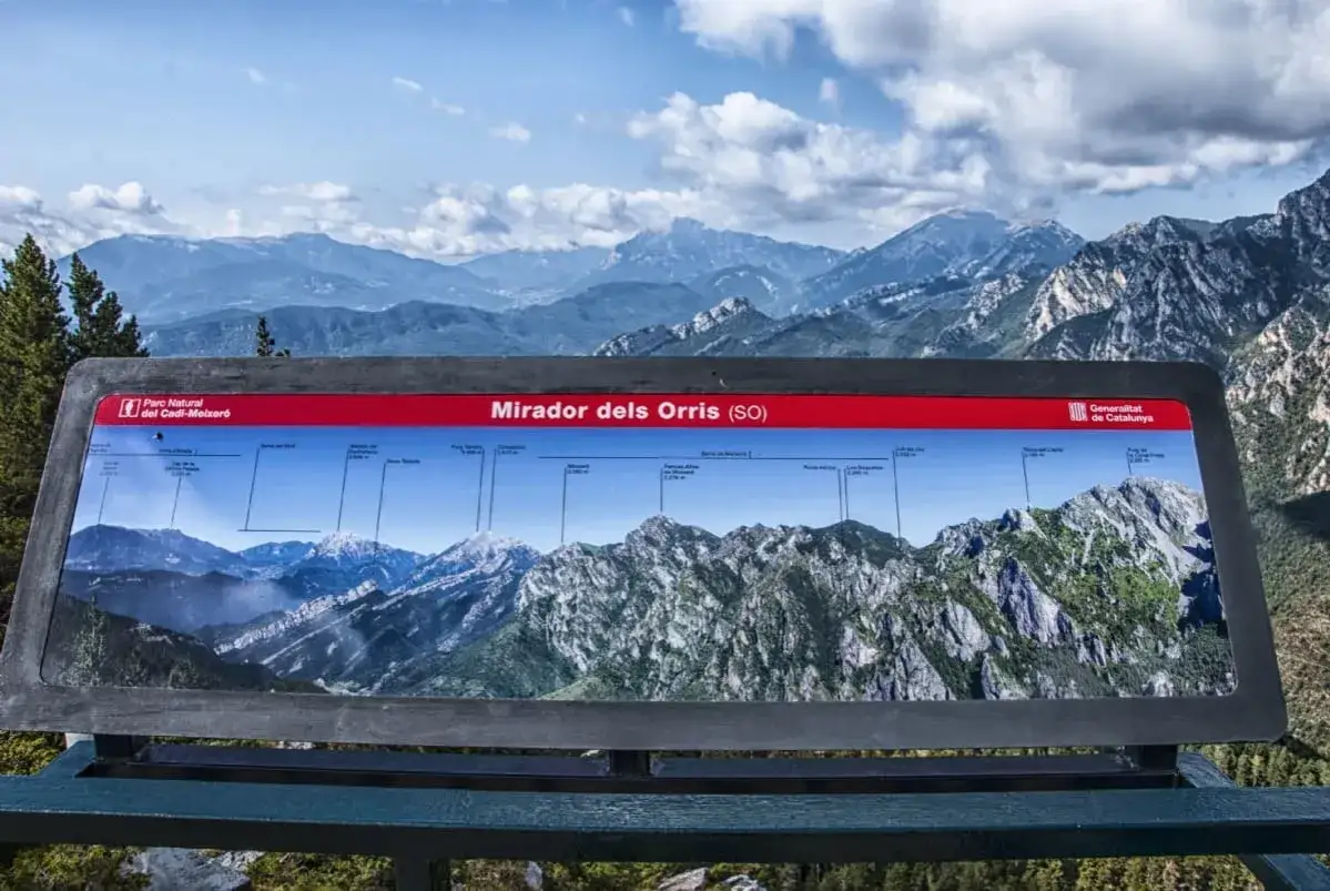 Panel informativo del **Mirador dels Orris** con vistas a majestuosas montañas nevadas y verdes bajo un cielo azul con nubes.