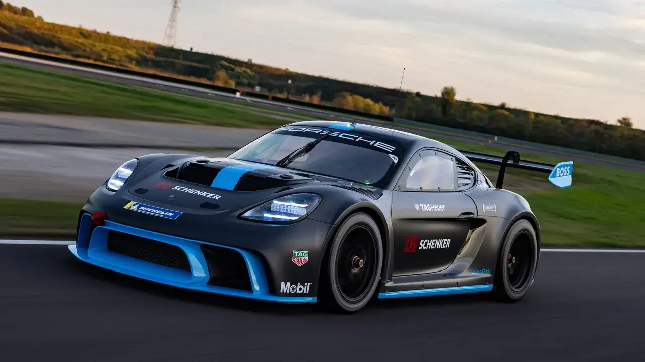 Czarny Porsche GT4 z niebieskimi pasami i logotypami DB Schenker i Michelin pędzi po torze wyścigowym.