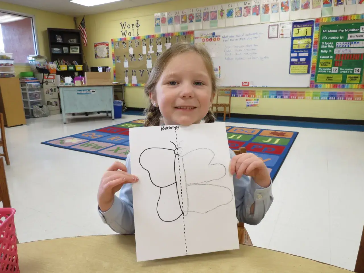 Ni&ntilde;a sonriente muestra dibujo de mariposa, aprendiendo sobre simetr&iacute;a primaria en clase.