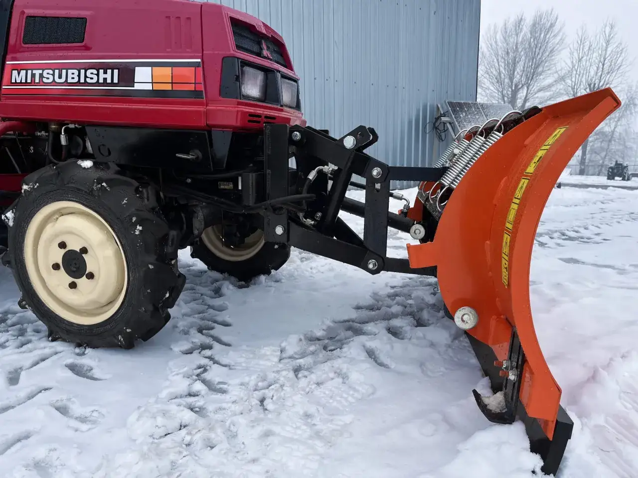 Czerwony traktorek Mitsubishi z zamontowanym pomarańczowym pługiem do śniegu do traktorka japońskiego, gotowy do pracy na zaśnieżonym podw&oacute;rku.