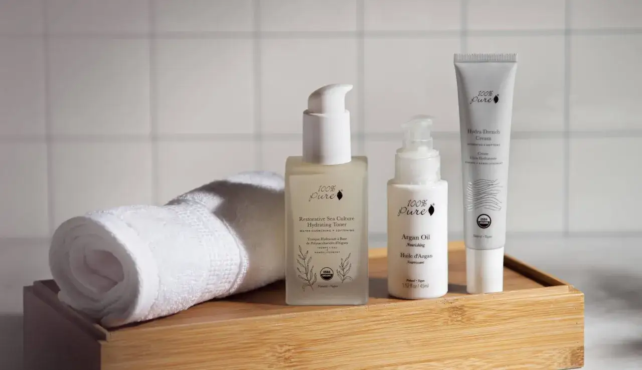 Hochwertige Kosmetik ohne Duftstoffe und Konservierungsmittel: Ein feuchtes Handtuch neben drei Pflegeprodukten von 100% Pure auf einem Holztablett.