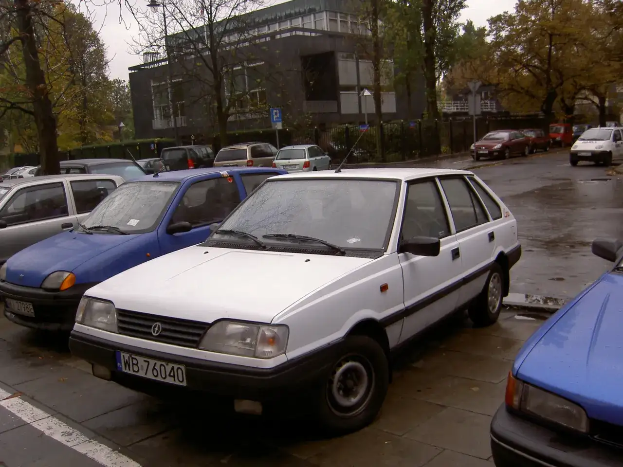 Biały Polonez Caro, który samochód jest w kole fortuny, stoi na mokrej ulicy obok niebieskiego Fiata Seicento.
