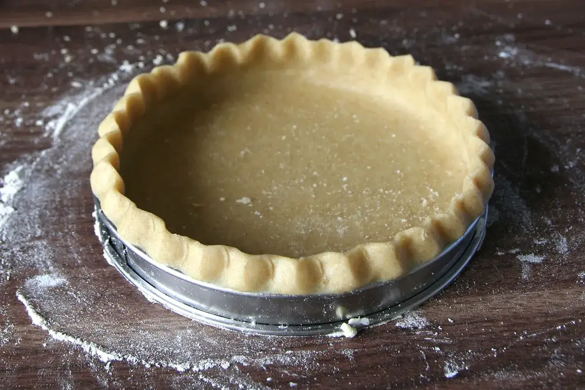 Une pâte à tarte maison, prête à être garnie, repose dans un moule à tarte sur un fond en bois parsemé de farine.