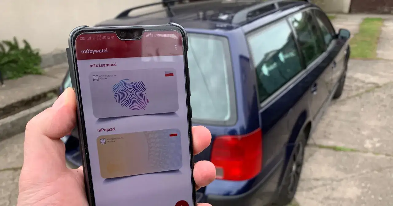 Na ekranie telefonu w aplikacji mObywatel widać cyfrowy dowód rejestracyjny pojazdu. Obok stoi ciemnoniebieski samochód.