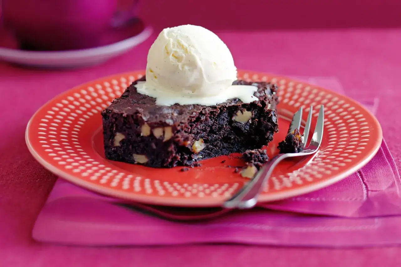 Ein Stück saftiger Brownie mit einer Kugel cremigem Vanilleeis, serviert auf einem roten Teller mit weißen Punkten. Ein Gabelrest liegt daneben.
