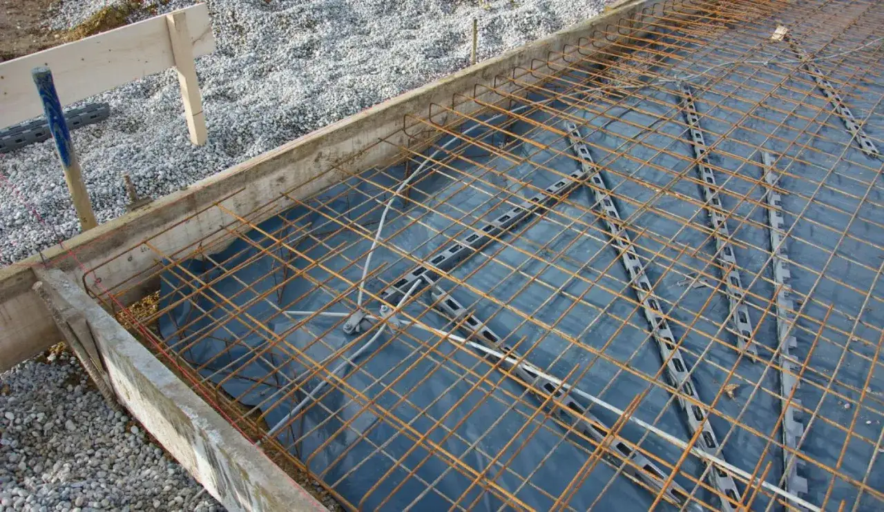 Préparation pour faire une dalle béton : coffrage en bois, film plastique, et treillis métallique rouillé. Gravier autour.