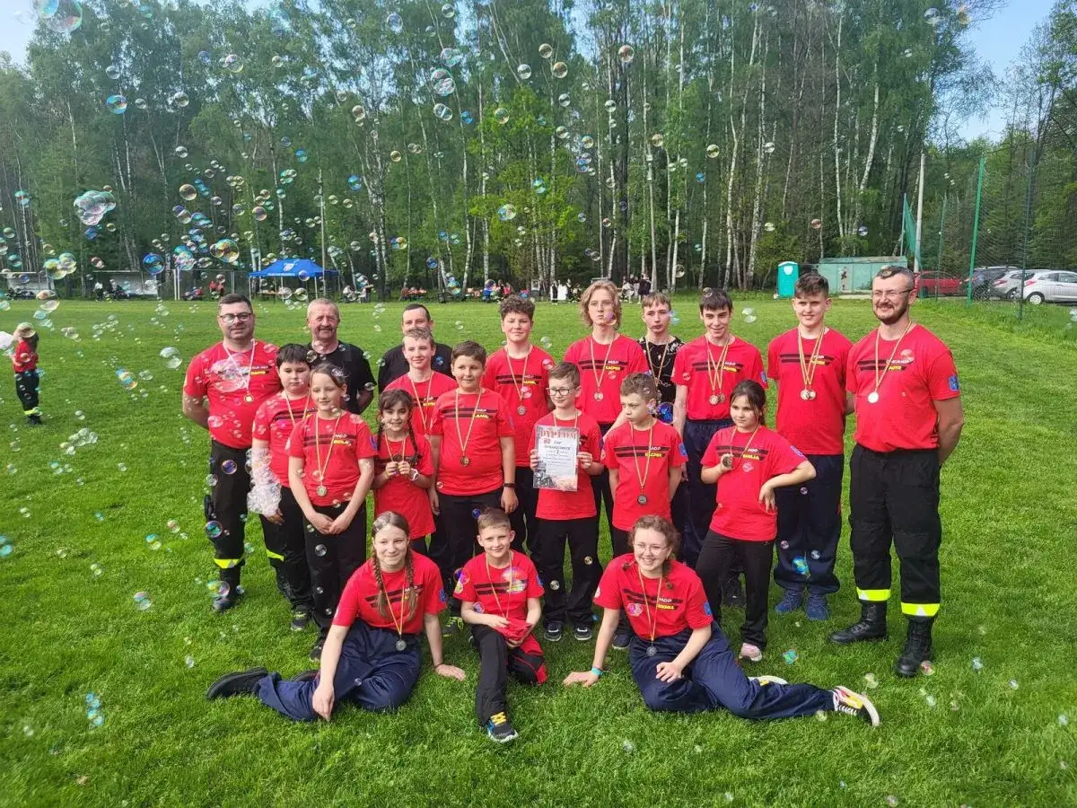 Uczestnicy zawodów CTIF w czerwonych koszulkach z medalami, otoczeni bańkami mydlanymi, pozują do zdjęcia na trawie.