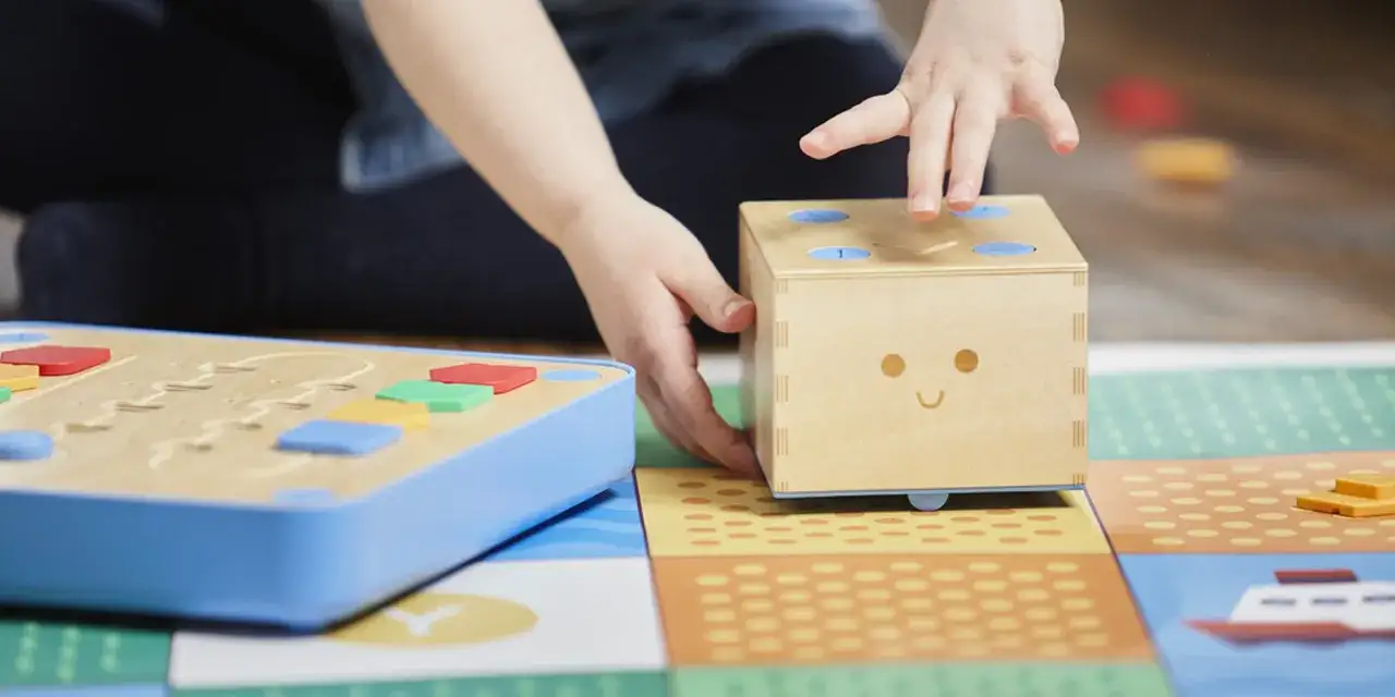 Bambino interagisce con un dado sorridente e blocchi colorati, esplorando i **giochi di programmazione** su un tappeto interattivo.