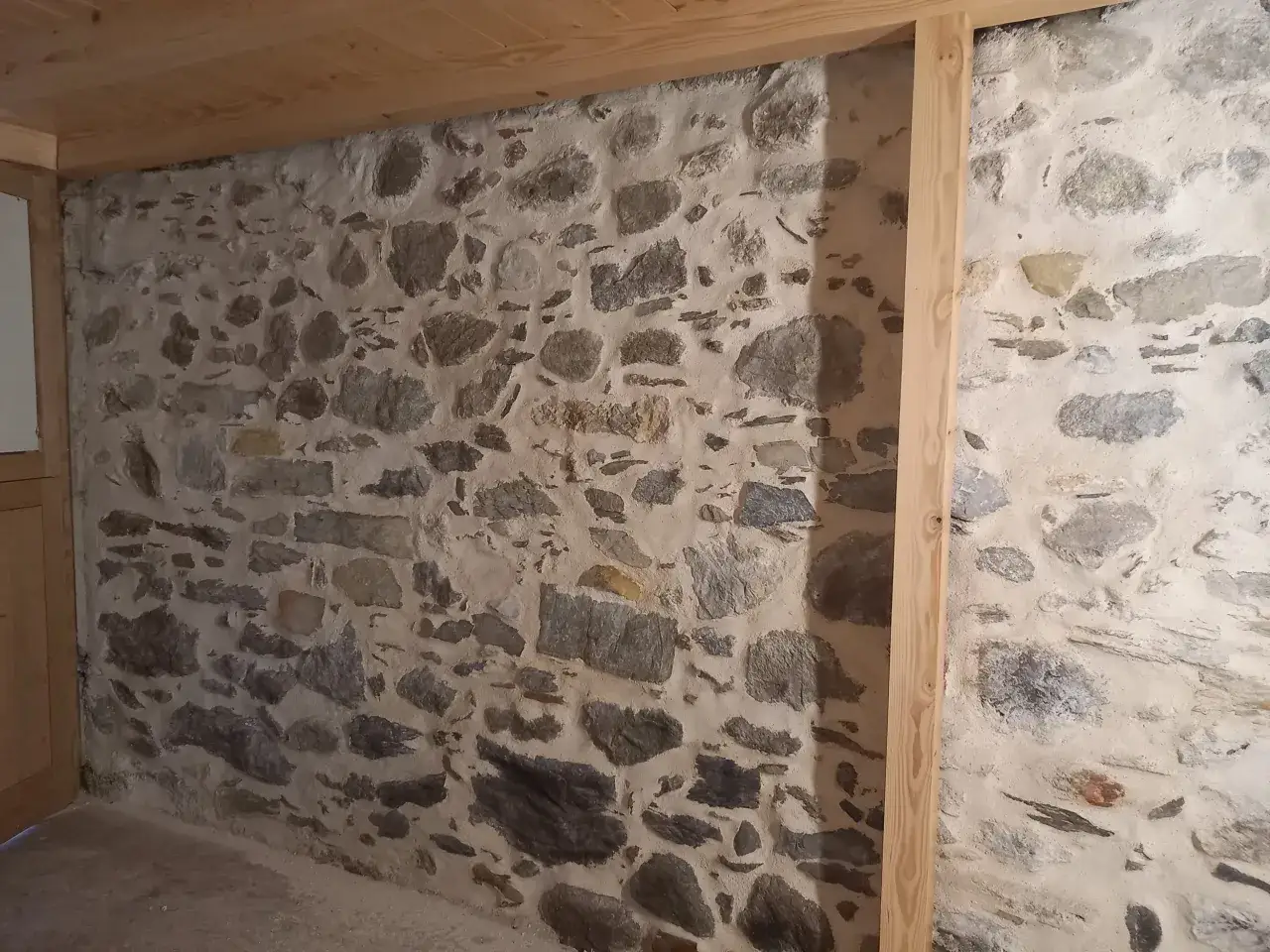 Mur en pierres brutes avec un joint pierre chaux traditionnel. Des poutres en bois clair encadrent la maçonnerie.